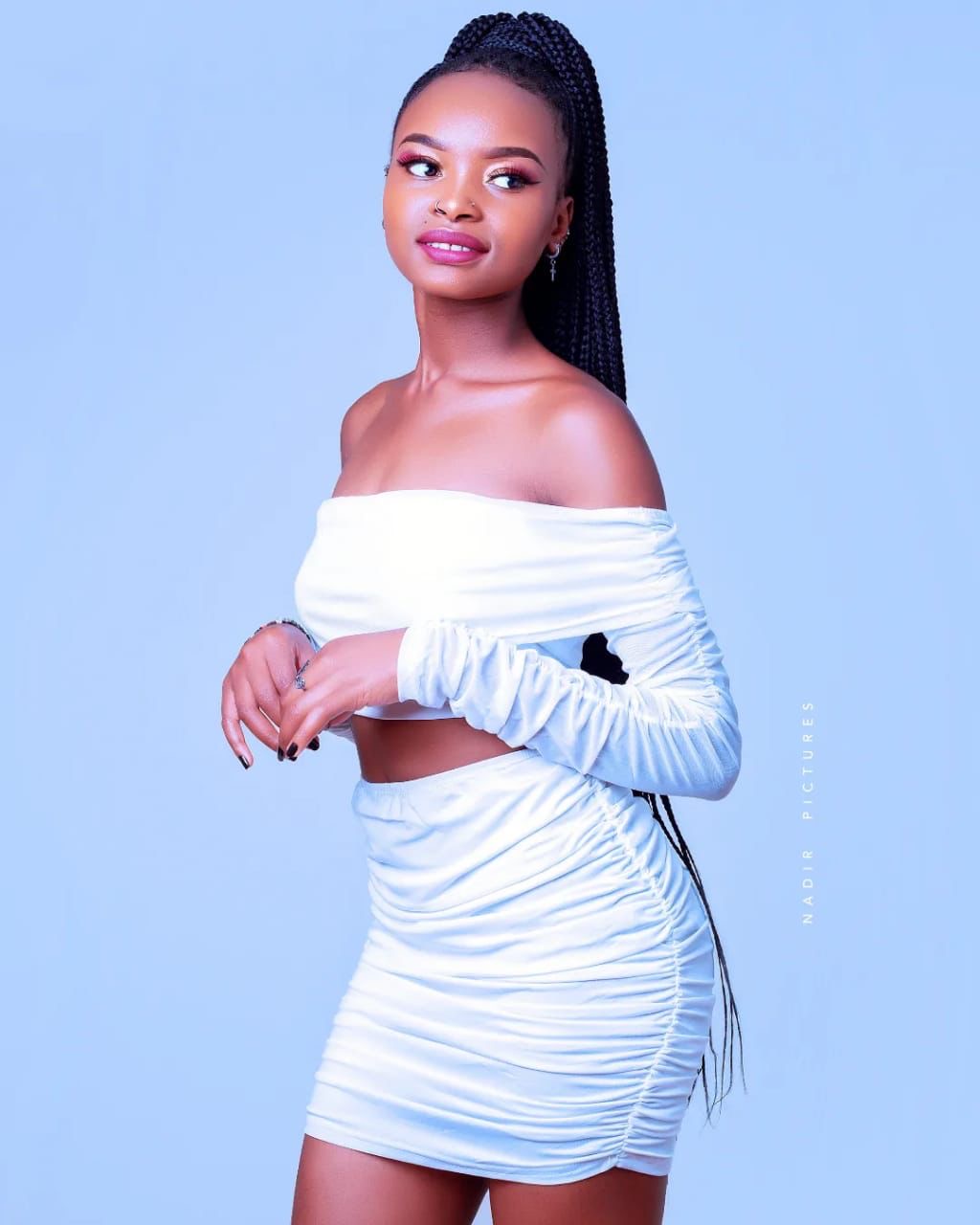 Breanda Naliaka, Senior Makeup Artist at She's Confident, ruched white satin two-piece with long box braids in a high ponytail, powder-blue backdrop