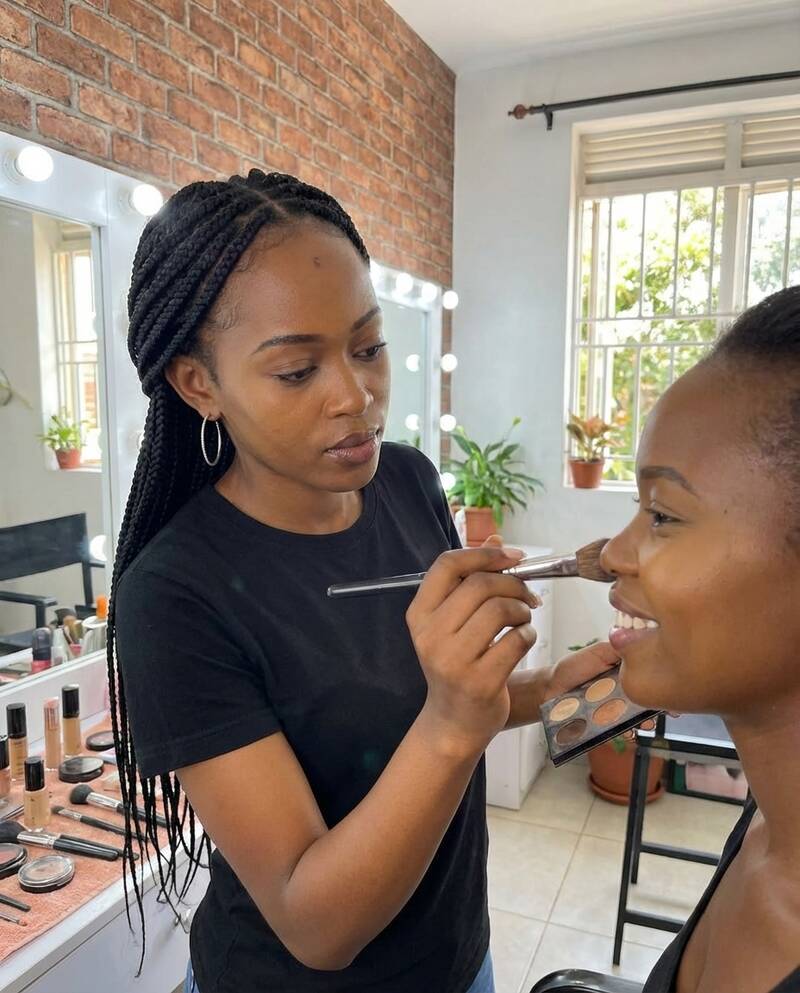 Stacey Njeri, Founder & Lead Artist at She's Confident, applying makeup to a client in a brick-walled studio with vanity mirror lights